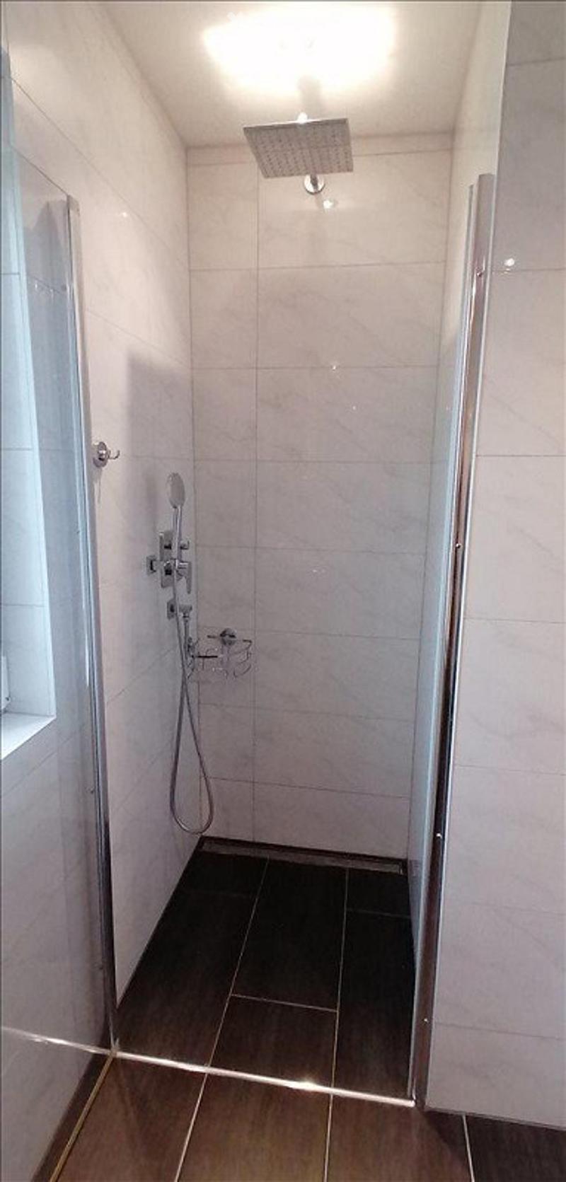 Modern, compact shower stall with white tiled walls, dark tiled floor, and a glass enclosure. Features a handheld showerhead and a small shower rack for storage.