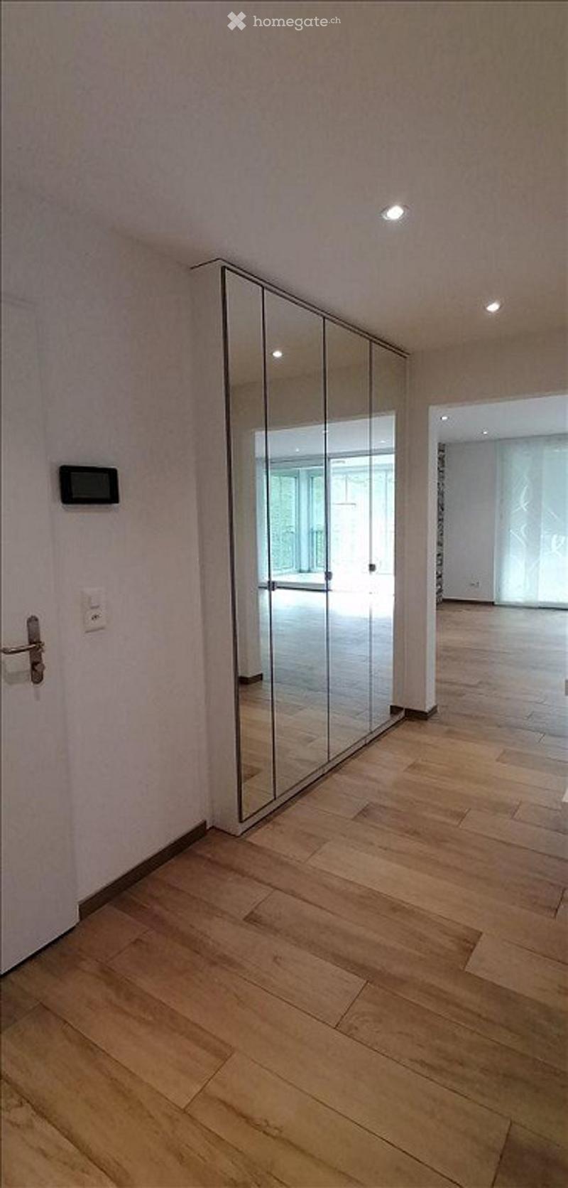 Empty room with light wooden flooring, white walls, and a large mirrored sliding door leading to another space.