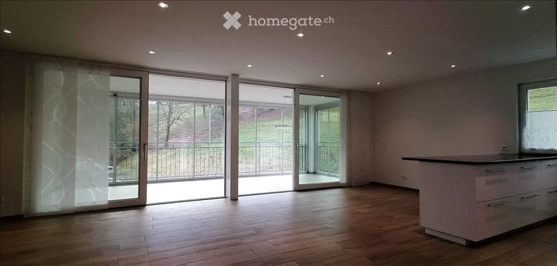 Empty room with wooden flooring, large sliding glass doors leading to a balcony, and a small kitchen counter visible on the right side.