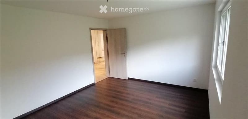 Empty room with wooden flooring, white walls, and a single window. An open doorway leads to another room.