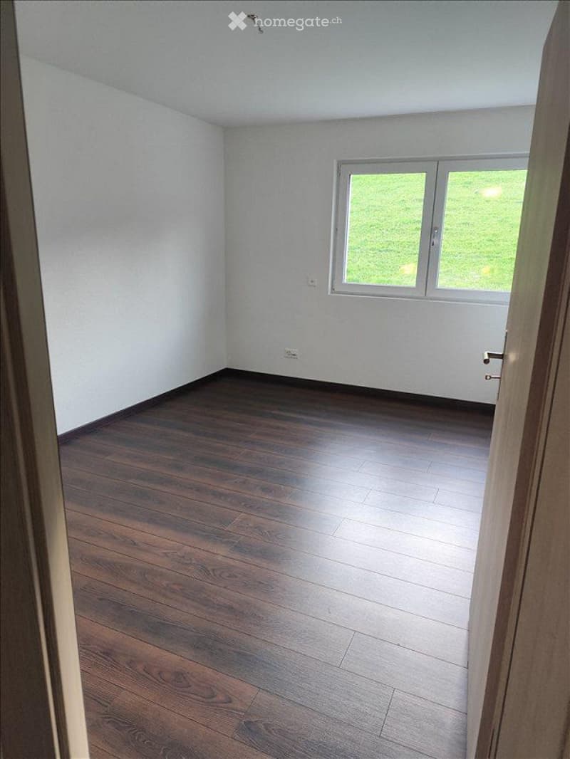 Empty room with wooden flooring and white walls, featuring a large window with white frames and a door visible on the right side.