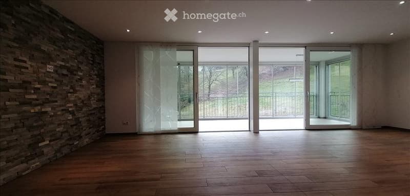 Empty room with wooden flooring, large glass sliding doors leading to a balcony, and a textured stone wall on one side.