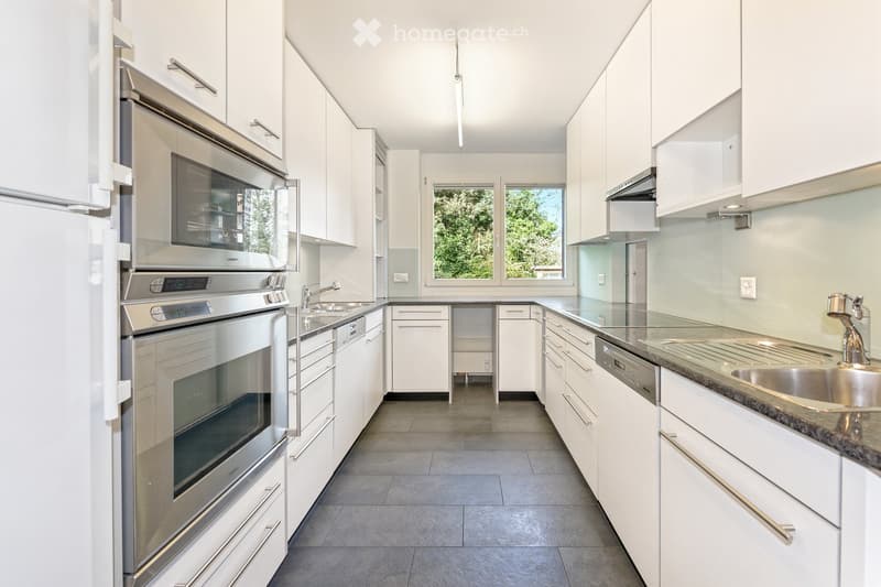 Modern kitchen with white cabinets, stainless steel appliances, granite countertops, and a large window overlooking greenery.