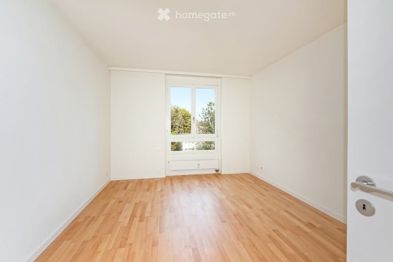 Empty room with light wooden flooring, white walls, and a large window with a view of trees outside.