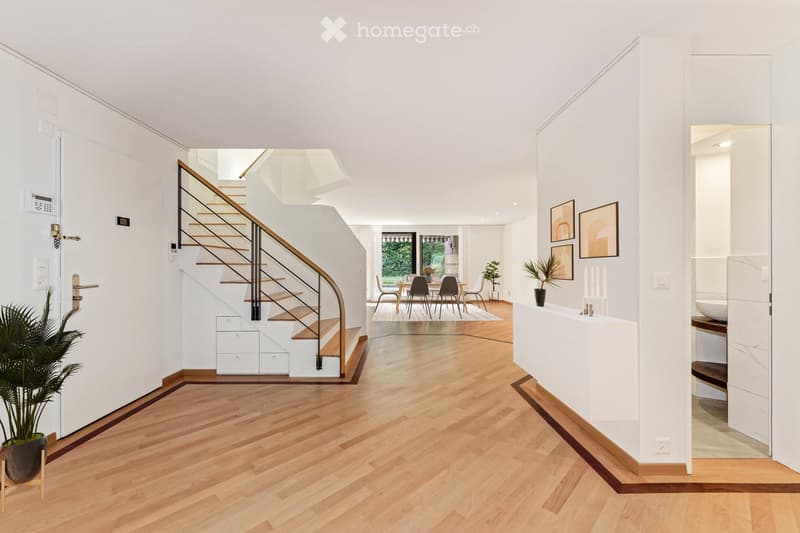 Bright, modern hallway with wooden flooring, white walls, and a staircase leading upwards. Features include a potted plant, a small console table with decorative items, and a dining area visible in the background.