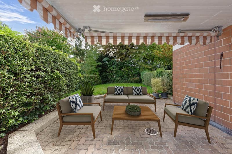 A well-maintained outdoor patio with a stone-paved floor, a covered awning, and a seating area featuring a sofa, chairs, and a small wooden table with a potted plant. The area is surrounded by lush greenery and brick walls.