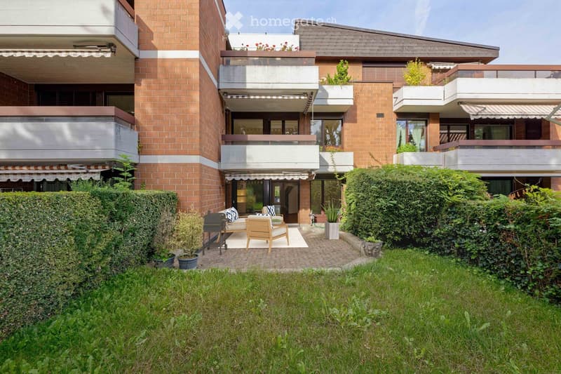 A modern multi-story building with balconies and a well-maintained garden area featuring a seating area with a table, chairs, and an umbrella.