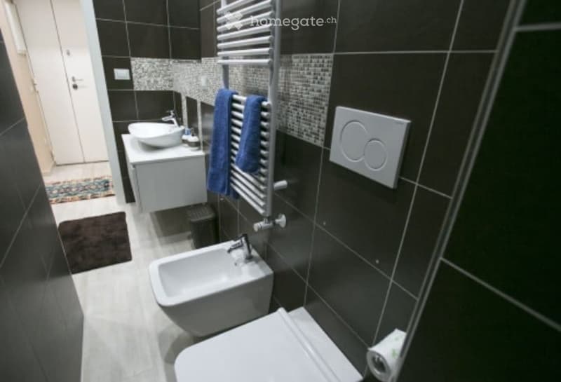 Modern bathroom with black tiled walls, white fixtures, a sink, toilet, bidet, towel, and a radiator.