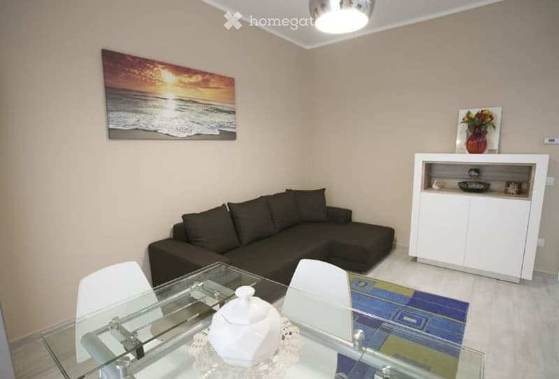 Modern living room with a dark sectional sofa, glass dining table with white chairs, white cabinet, and decorative artwork on the walls.