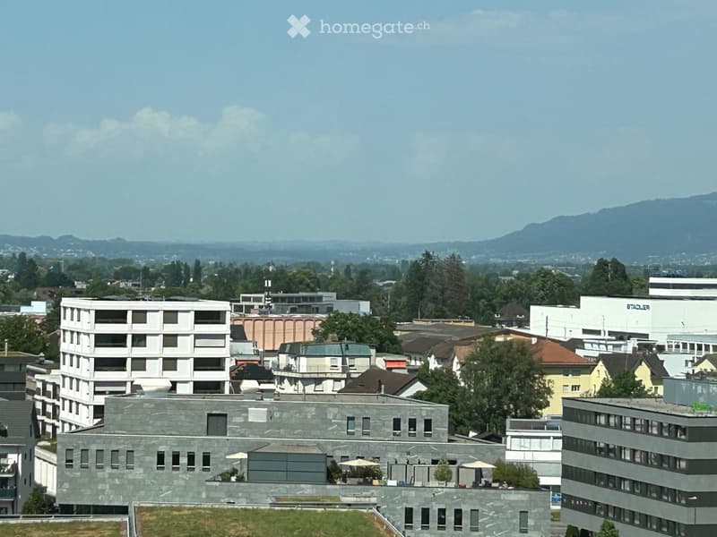 A modern multi-story building complex with a flat roof, surrounded by other structures and greenery, with a scenic mountain backdrop.