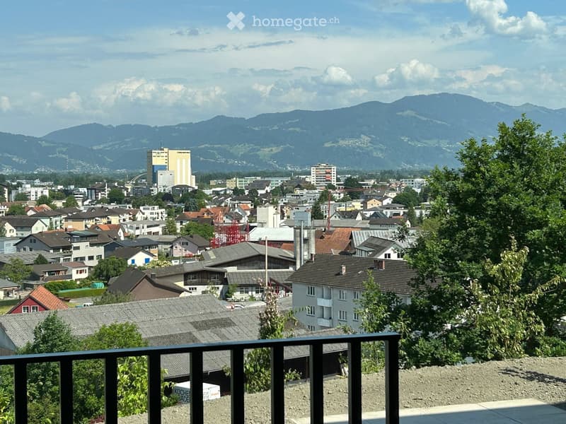 A balcony overlooking a town with houses, trees, and distant mountains under a partly cloudy sky.