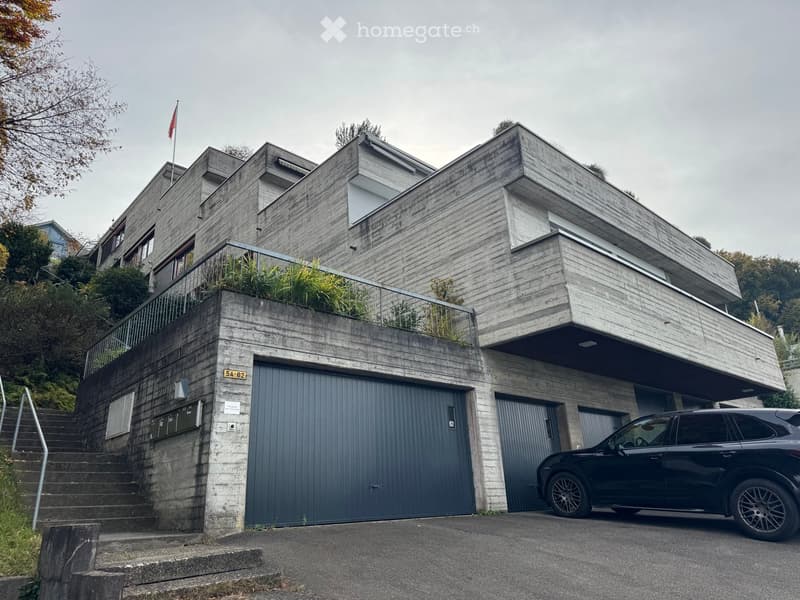 Modern two-story house with concrete and wood exterior, featuring a large garage, balcony, and a parked SUV.