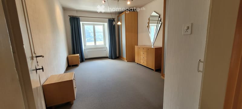 A furnished room with a wardrobe, dresser, mirror, and curtains. The room has a carpeted floor and a window with a radiator below it.