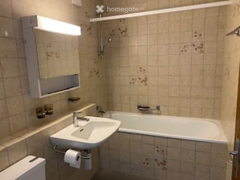 Bathroom with beige tiled walls and floor, a white sink with a mirror above it, a bathtub, and a toilet. The lighting is bright, and the space appears clean and functional.
