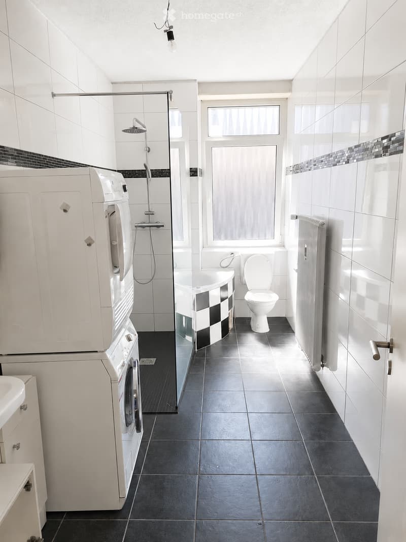 Modern bathroom with white tiles, black accents, a bathtub, toilet, shower, washing machine, and radiator.