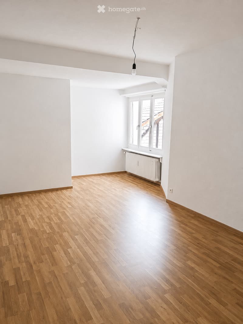 Empty room with wooden flooring, white walls, and a large window with a radiator below it. The room is well-lit with natural light and has a simple, clean design.