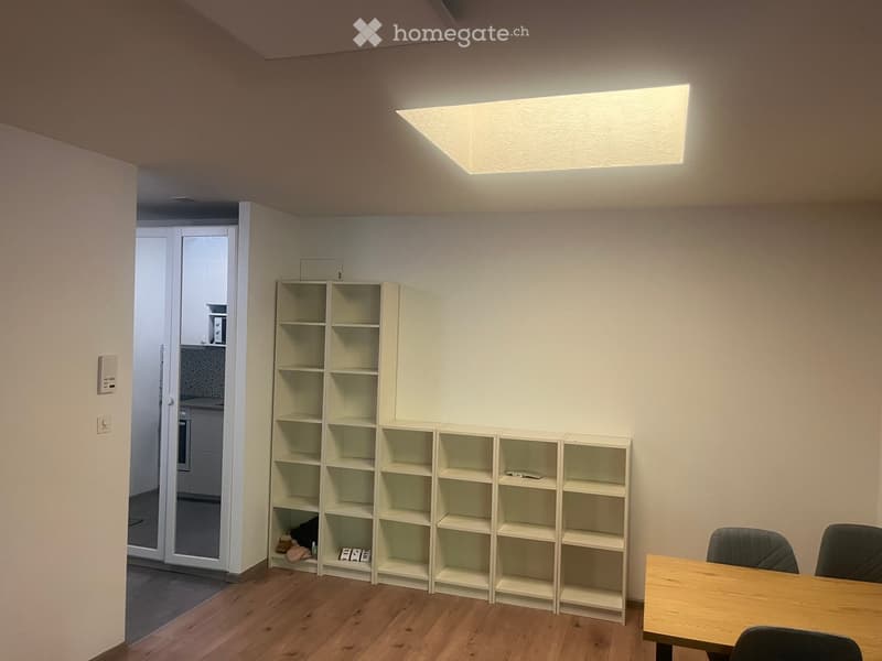 A small, unfurnished room with a wooden floor, white walls, and a large rectangular light fixture on the ceiling. A wooden table with chairs is positioned near the right side, and a white shelving unit is against the left wall.