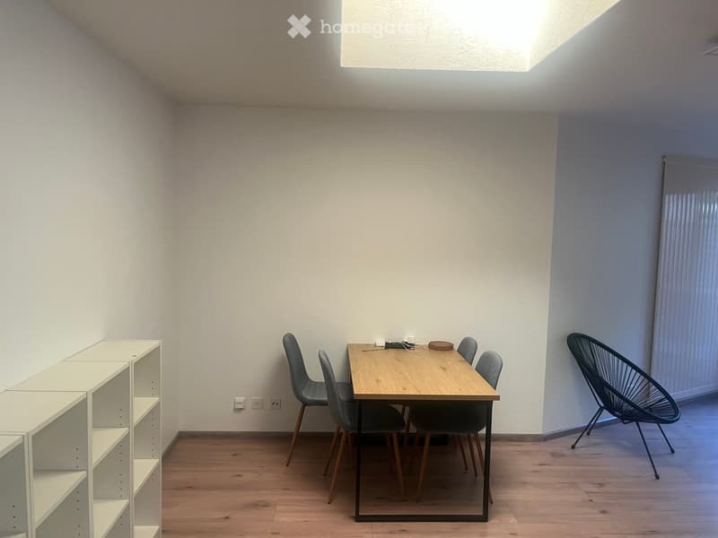 A modern, minimalist room with light wooden flooring, white walls, and a skylight. It features a wooden dining table with four chairs, a sleek white shelving unit, and a contemporary black wire chair.