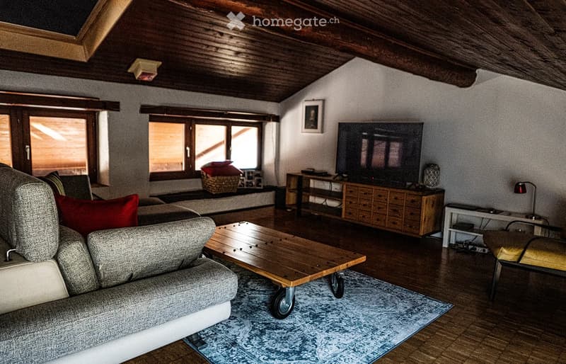 A cozy living room with wooden beams on the ceiling, large windows, a grey sofa with red cushions, a wooden coffee table, a television on a wooden stand, and a small side table with a lamp.