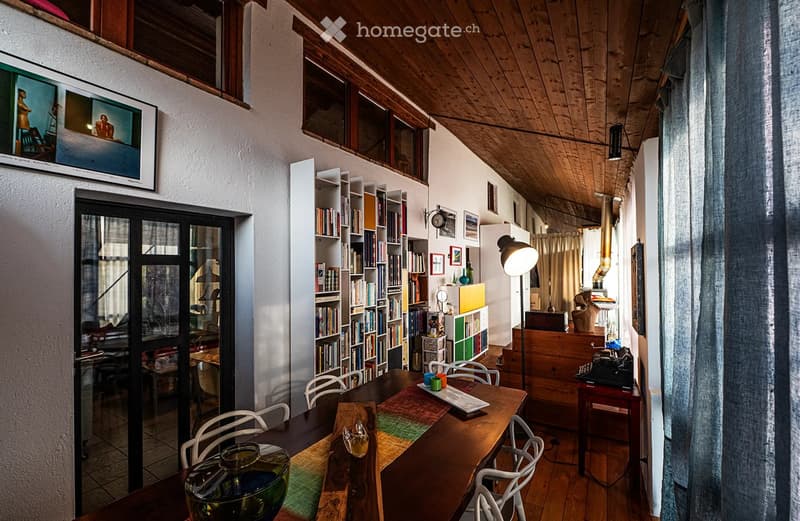 A cozy, well-lit room with wooden floors and exposed wooden beams on the ceiling. Features include a large wooden dining table with chairs, a bookshelf filled with books, a floor lamp, a television mounted on the wall, and a curtained window. The room als