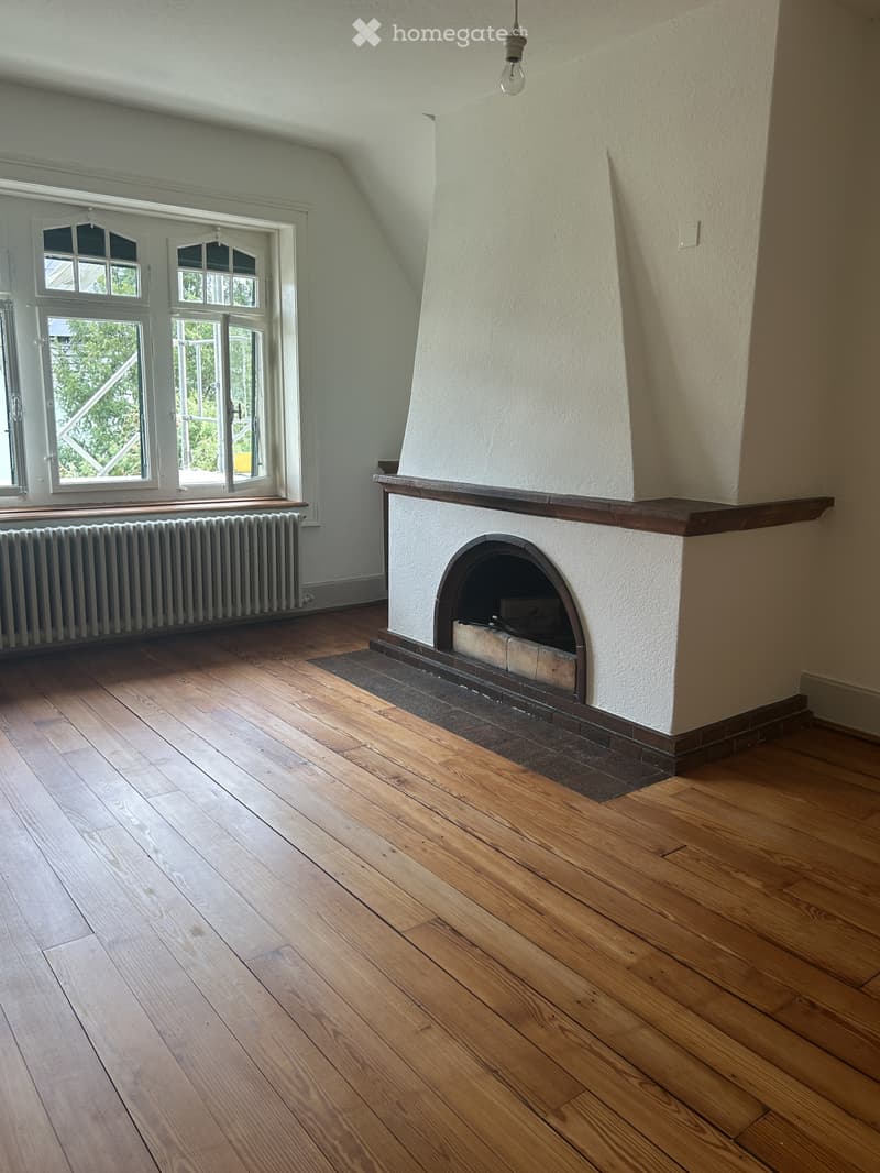 Empty room with wooden flooring, white walls, fireplace, radiator, and large windows with double doors leading outside.