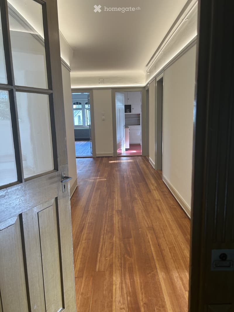 A long, narrow hallway with wooden flooring, light-colored walls, and recessed lighting. The hallway features double doors at the far end leading to another room and a partially visible kitchen area.