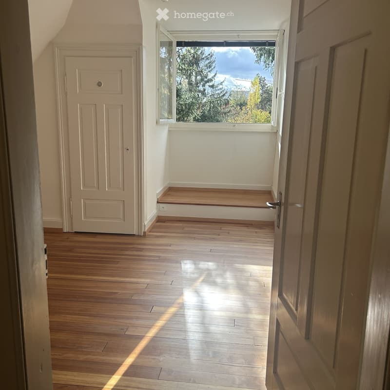 Empty hallway with wooden flooring, white walls, and a large window letting in natural light. A closed door is visible on the left side.