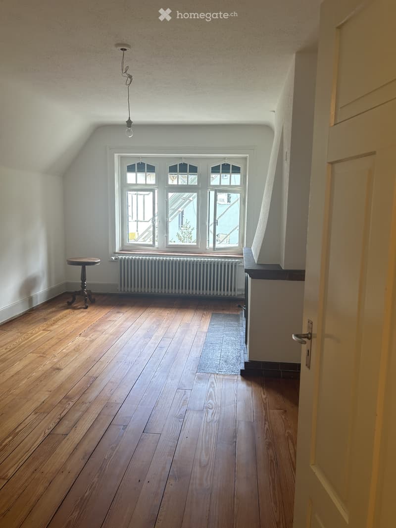 Empty attic room with wooden flooring, large windows, radiator, and a small wooden stool near the window.