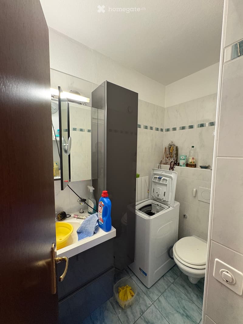 A compact bathroom with a toilet, washing machine, sink, mirror, and various personal care items. The space features tiled walls and a small shelf holding bottles and other items.