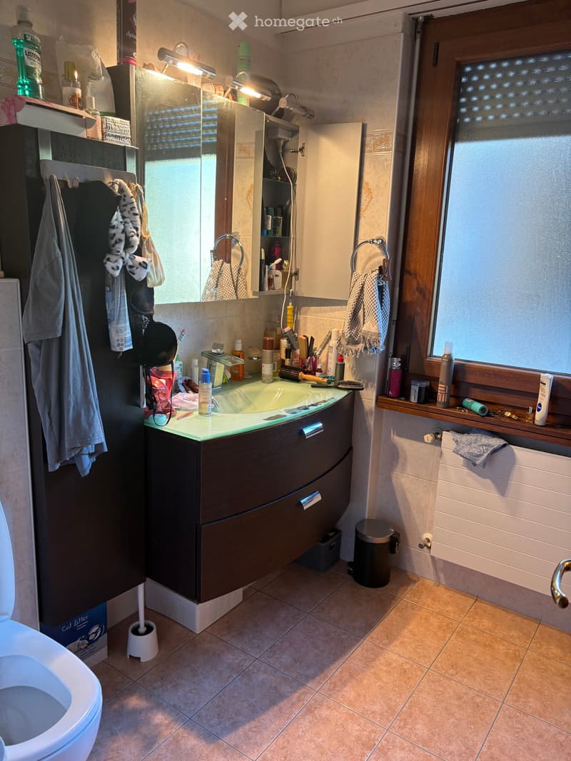 Modern bathroom with a sink, mirror, cabinet, and toilet. The sink has various toiletries, and there are towels and a trash bin visible.