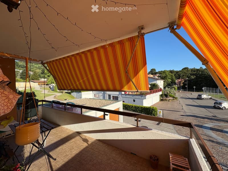 Balcony with wooden decking, metal railing, and striped awnings, overlooking a paved area with parked cars and greenery.