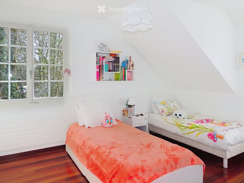 Cozy children's bedroom with two single beds, colorful bedding, a nightstand with a lamp, a bookshelf filled with books, and decorative toys.