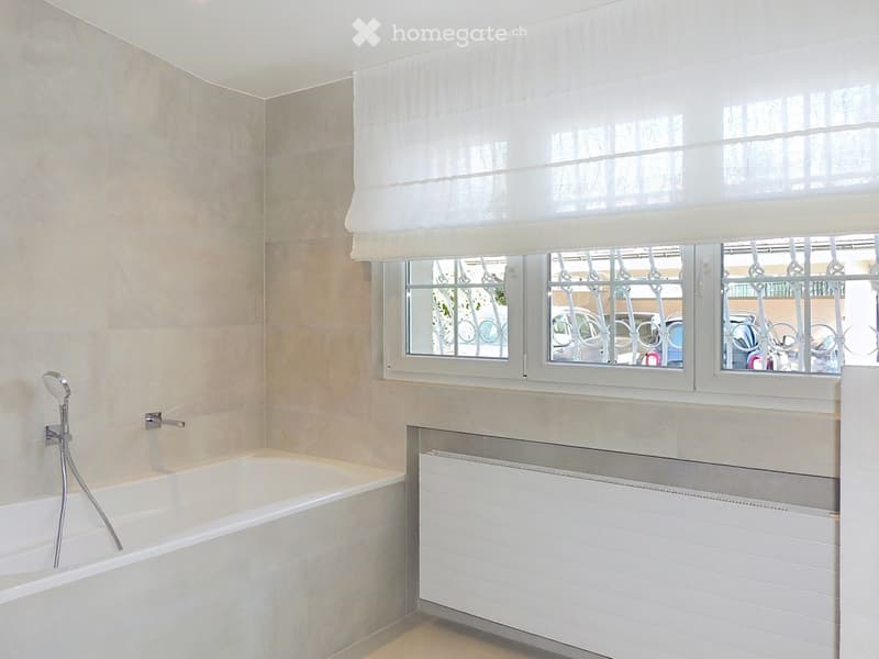 Bathroom with a bathtub, showerhead, radiator, and a window with white curtains and metal bars.