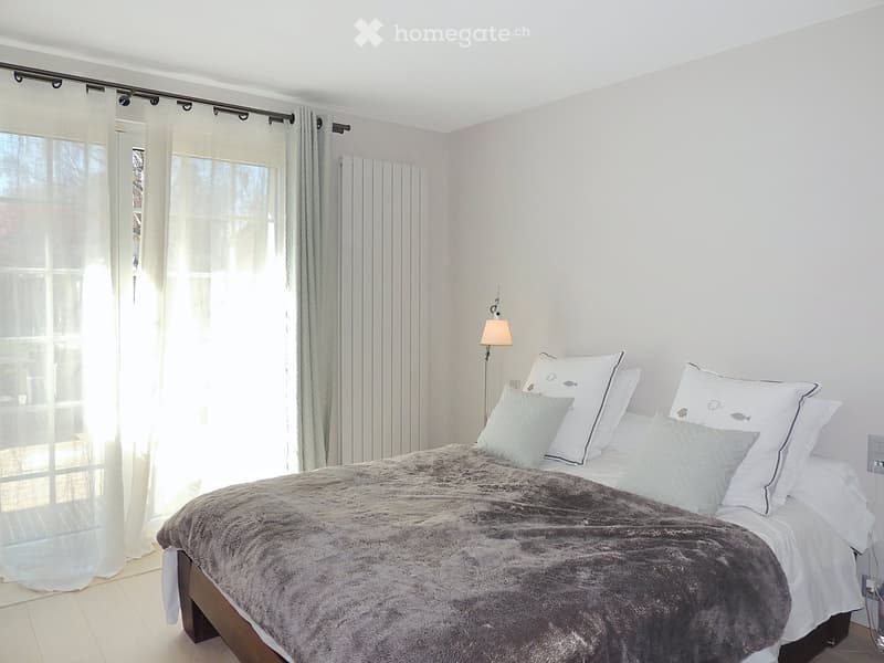 Bright bedroom with a large bed, multiple pillows, a fur throw, and a wall-mounted lamp. Large windows with sheer curtains allow natural light to fill the space.