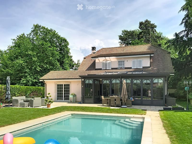 A two-story house with a tiled roof and light pink facade, surrounded by a well-maintained garden with a swimming pool, patio furniture, and outdoor seating area.