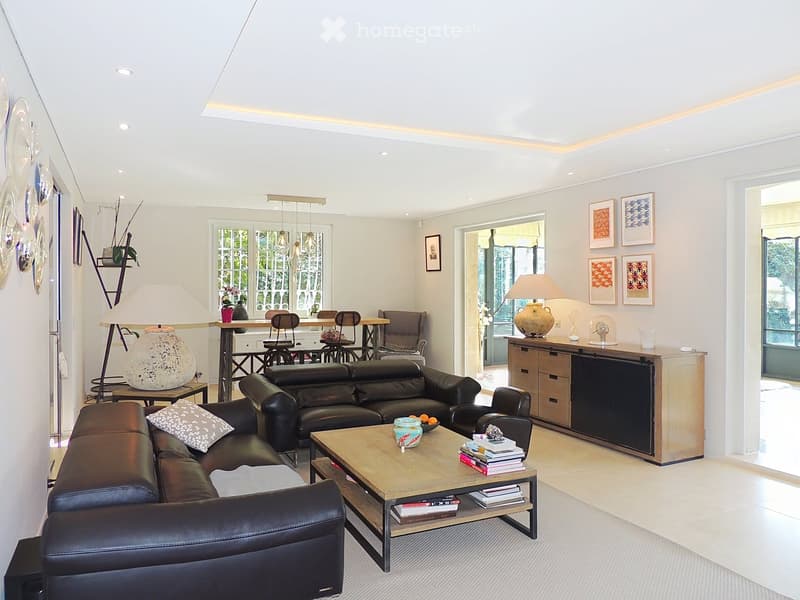 Modern living room with a black leather sofa, wooden coffee table, dining area with chairs, decorative lamps, and artwork on the walls.