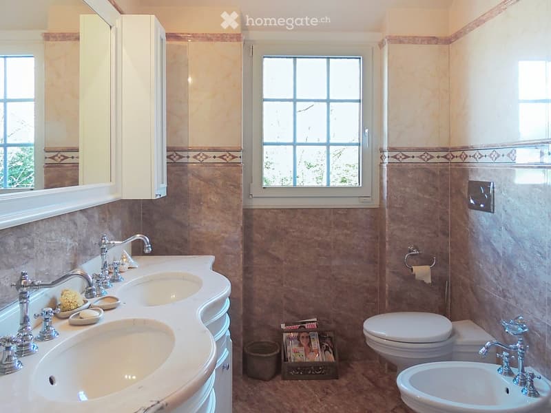 Elegant bathroom with double sink vanity, chrome faucets, toilet, bidet, and decorative cabinetry. Features large windows and warm tilework.