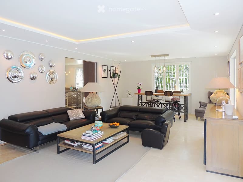 Modern living room with a black leather sectional sofa, wooden coffee table, dining area with chairs, and decorative wall plates.