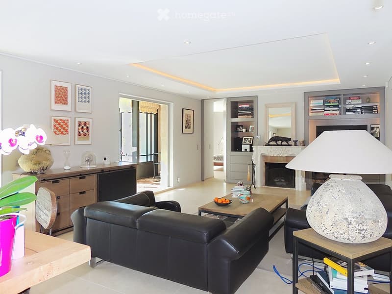Modern living room with white walls, a black leather sofa, a wooden coffee table, a side table with a lamp, a fireplace, and a bookshelf. The room features large windows and a view of the outdoor area.