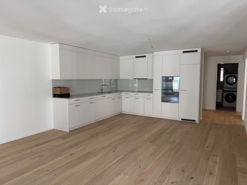 Modern kitchen with white cabinets, wooden flooring, and stainless steel appliances. A sink and faucet are visible, along with an oven. A small box is placed on the counter.