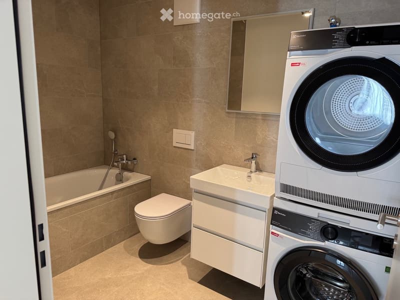Modern bathroom with beige tiles, a bathtub, toilet, sink, and a stacked washer/dryer unit.