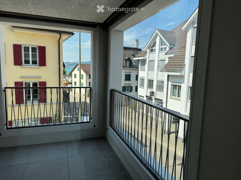 Balcony with tiled floor, metal railing, overlooking a street with buildings and a distant view of a body of water.