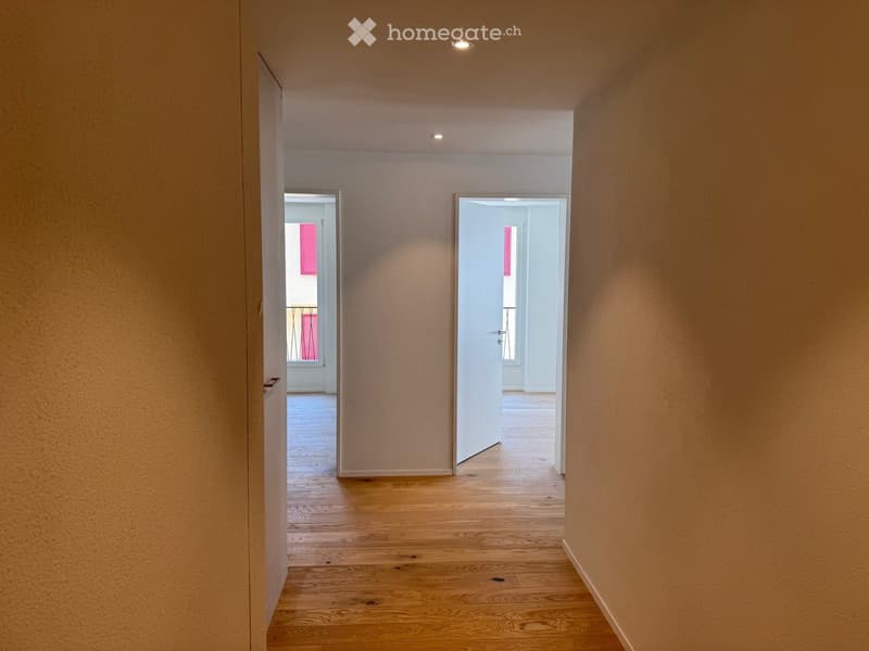 Empty hallway with wooden flooring, white walls, and recessed ceiling lights. Two open doors lead to adjacent rooms.