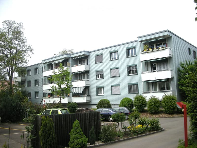 A modern, multi-story apartment building with light blue exterior walls, white balconies, and multiple windows. The building is surrounded by greenery and has several parked cars in the foreground.