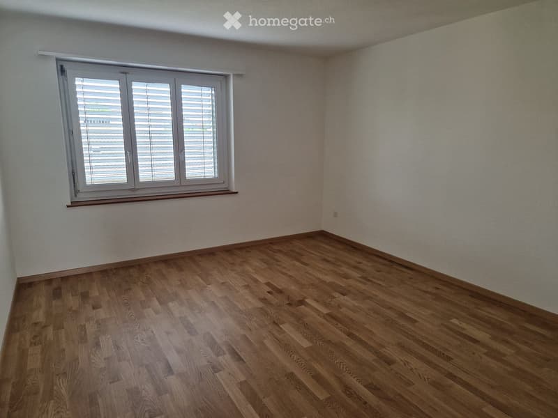 Empty room with wooden flooring and white walls, featuring a large window with blinds and a small wooden sill.