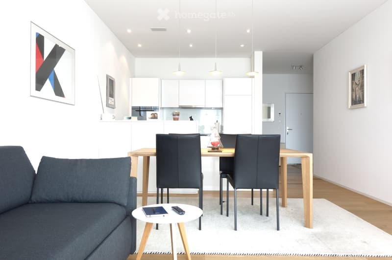 Modern, open-plan living and dining area with a grey sofa, coffee table, dining table with chairs, and a kitchen area with white cabinets and appliances.