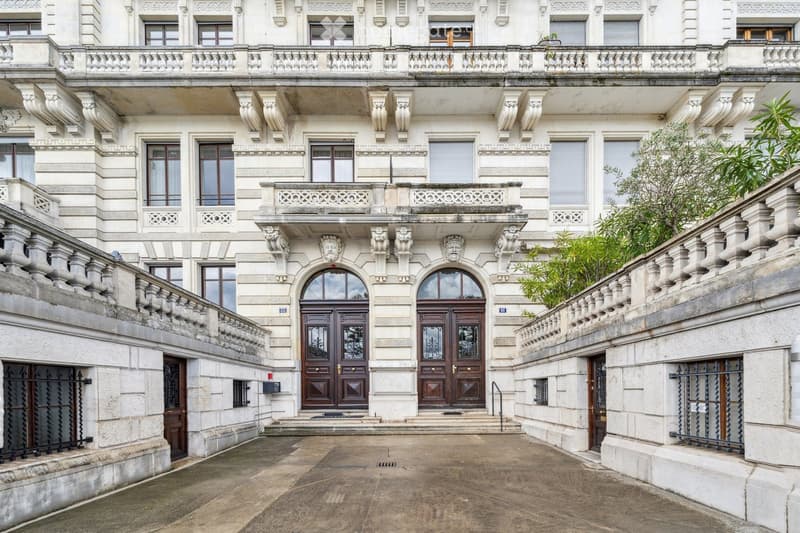 Grand classical building with ornate stone facade, large arched entrance doors, decorative balustrades, and multiple windows.