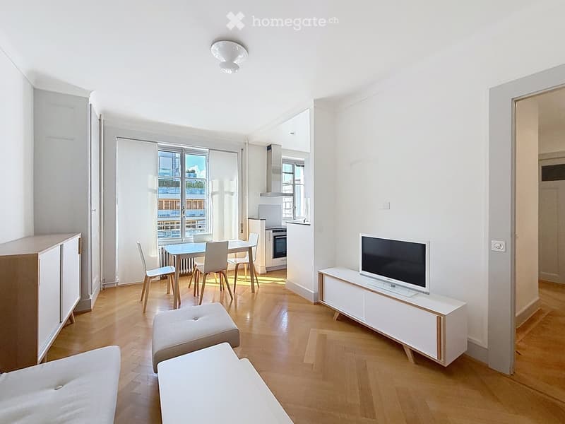 Bright, modern living and dining area with wooden flooring, white walls, and large windows offering a view. Features include a sofa, coffee table, TV stand, dining table with chairs, and a small kitchen area with cabinets and an oven.