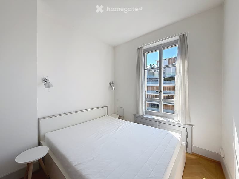 Minimalist bedroom with a single bed, white bedding, a small round stool, and wall-mounted lamps. The room features light-colored walls, wooden flooring, and a large window with curtains.