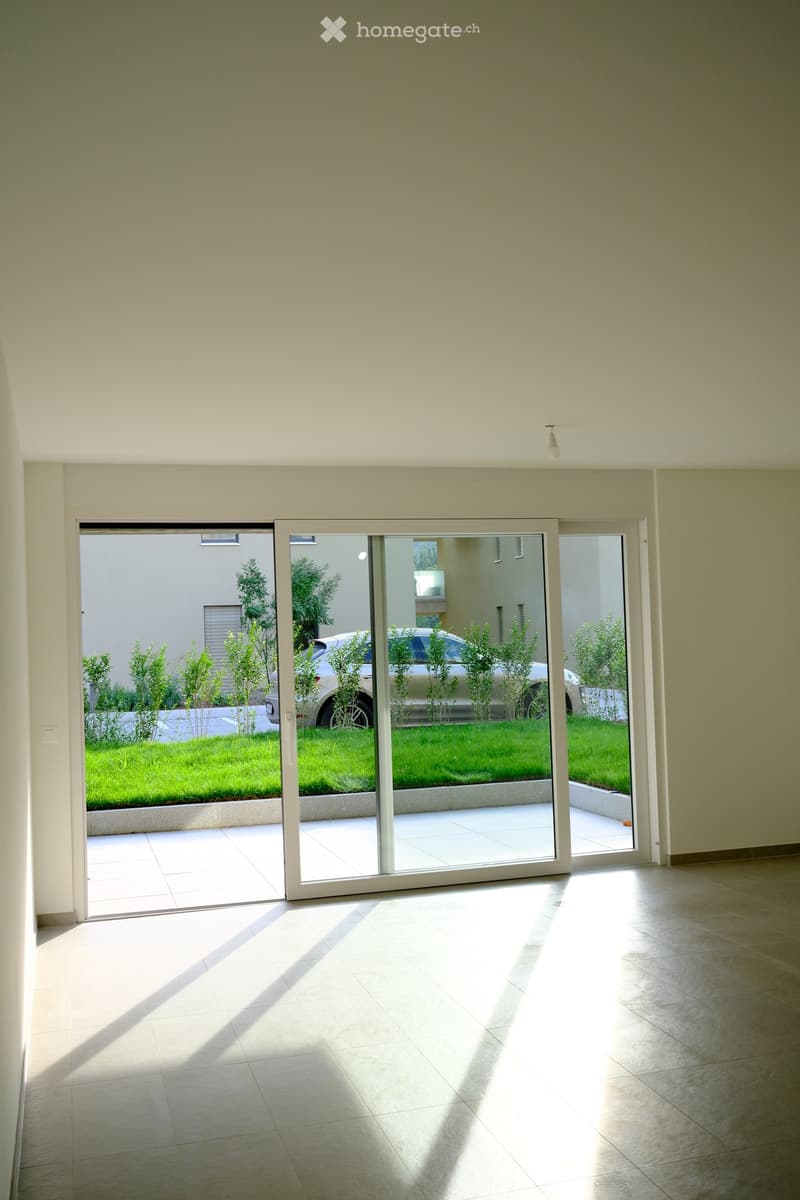 Bright, empty room with large sliding glass doors leading to a garden area. The room has tiled flooring and white walls, with natural light streaming in.